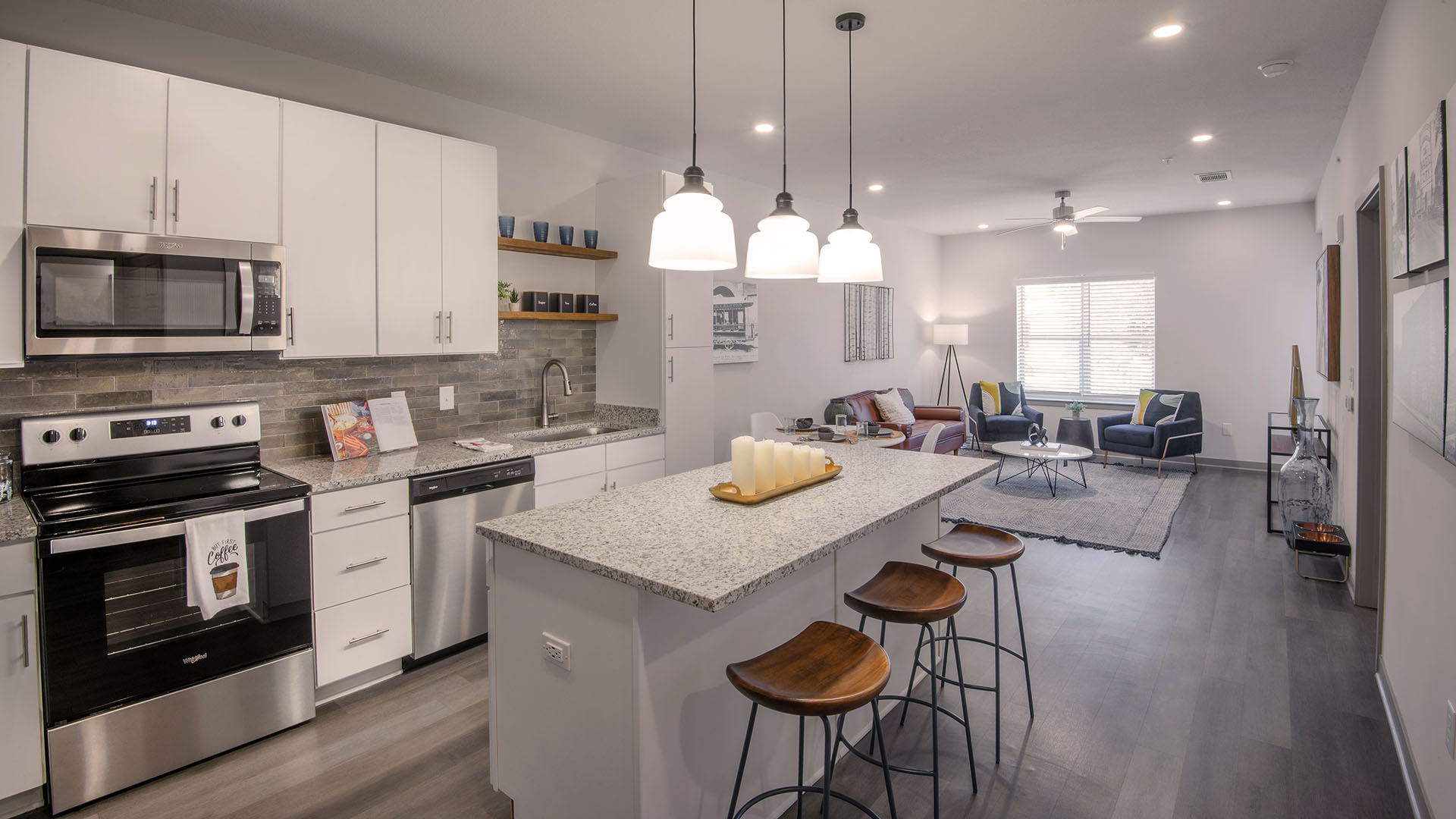 birmingham al apartments - Axel Row Apartments - Bright kitchen adjacent to living room featuring stainless steel appliances, white cabinetry, granite countertop island with wooden stool seats, and three white hanging ceiling lights.