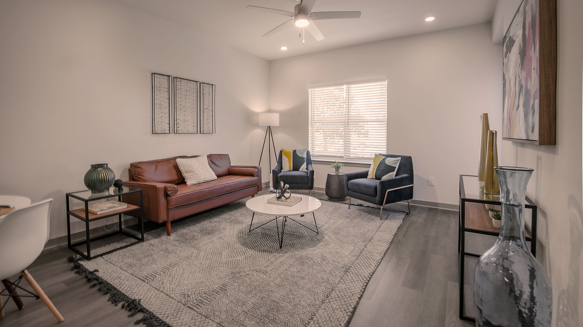 apartments in birmingham for rent - Axel Row Apartments - Modern livingroom featuring brown leather sofa, blue suede chairs, circular coffee table, white walls accented with artwork, ceiling fan with lights, and large window with shades
